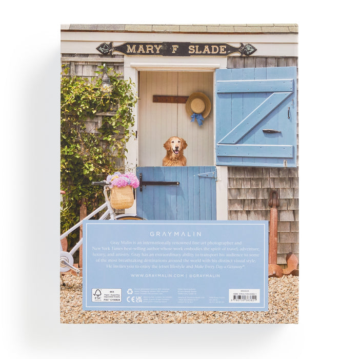Gray Malin’s Nantucket 1000 Piece Book Box Puzzle features a golden retriever at a rustic shed with a blue door labeled Mary F Slade, and a vintage bicycle with flowers—capturing the brands signature charm.