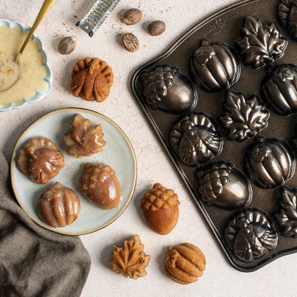 Acorn-shaped metal mold tray filled with baked acorn cookies, alongside plated acorn cookies and scattered acorns on a light stone surface.