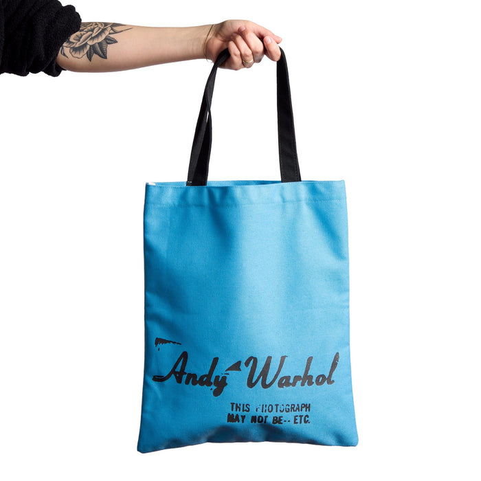 A person with a tattooed forearm holds up the Warhol Soup Can Volume 3 Canvas Tote Bag by Andy Warhol Foundation For The Visual Artsโa light blue tote with black handles and text on a white background.