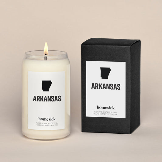 A lit Arkansas Homesick candle displayed next to its boxed packaging on a dark cream background.