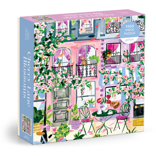 Sabina Fenn's Cherry Lane Blossoms 1000 Piece Puzzle features a vibrant café scene with pink blossom trees, outdoor seating, and charming balconies—perfect for puzzle enthusiasts.