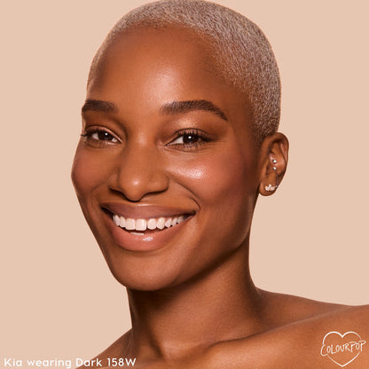 Model with close-cropped platinum hair smiles, showcasing ColourPop Pretty Fresh Creamy Concealer in shade Dark 158 W with smooth, radiant coverage; tight headshot on a soft beige background.