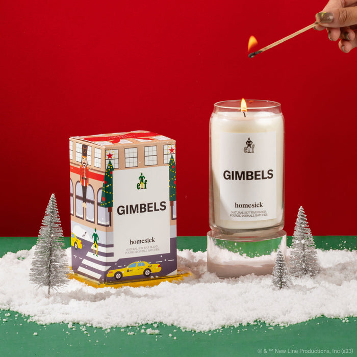 A hand coming in to light the Gimbels candle. Next to the candle is its packaging. Both are shot in a holiday environment on a green surface, with fake snow flakes, and a red background.