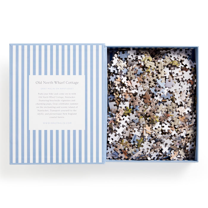 A partially open blue and white striped box reveals the Gray Malin Nantucket 1000 Piece Book Box Puzzle. The lid features info about Old North Wharf Cottage, inspired by stunning Nantucket photography. Brand: Gray Malin.