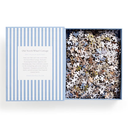 A partially open blue and white striped box reveals the Gray Malin Nantucket 1000 Piece Book Box Puzzle. The lid features info about Old North Wharf Cottage, inspired by stunning Nantucket photography. Brand: Gray Malin.