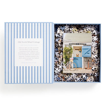 A blue and white striped Gray Malin Nantucket 1000 Piece Book Box Puzzle by Gray Malin, partially open, reveals a seaside cottage scene with blue doors, a pink bicycle, and a golden retriever. A description is printed inside the box lid.