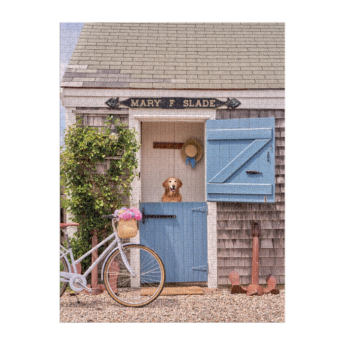 A brown and white dog stands in a Dutch doorway of a small wooden building, with a bike and flowers nearby—captured in the Gray Malin Nantucket 1000 Piece Book Box Puzzle by Gray Malin.