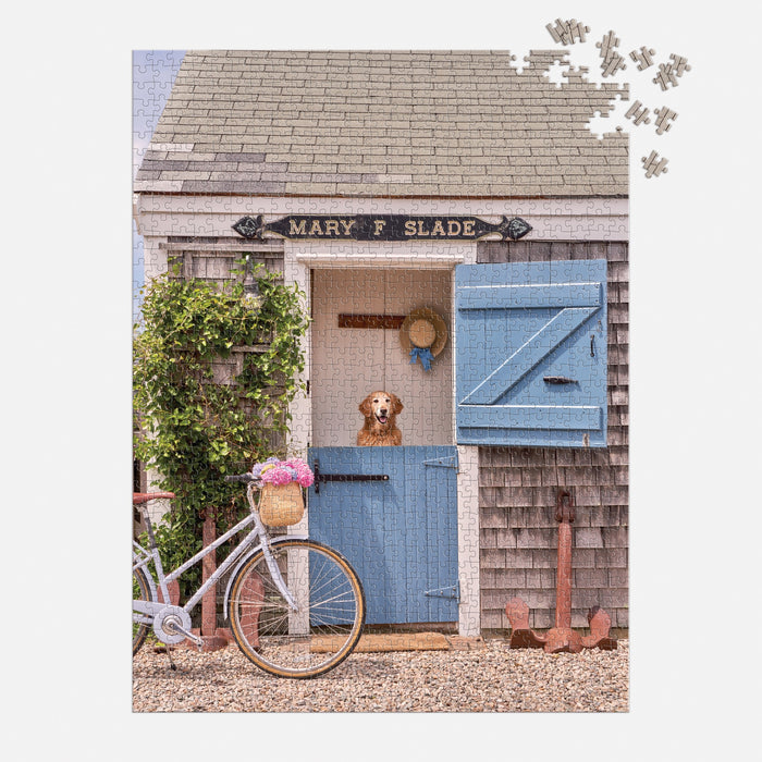 The Gray Malin Nantucket 1000 Piece Book Box Puzzle by Gray Malin features a brown and white dog at the blue door of a shingled cottage, with a white bicycle, flowers, and a Mary F. Slade sign completing the picturesque scene.