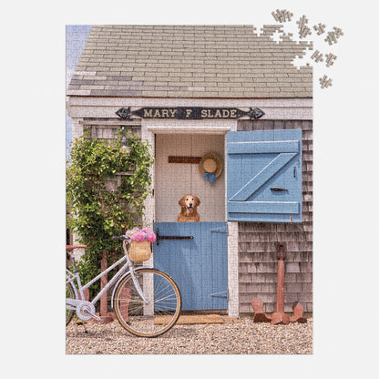 The Gray Malin Nantucket 1000 Piece Book Box Puzzle by Gray Malin features a brown and white dog at the blue door of a shingled cottage, with a white bicycle, flowers, and a Mary F. Slade sign completing the picturesque scene.