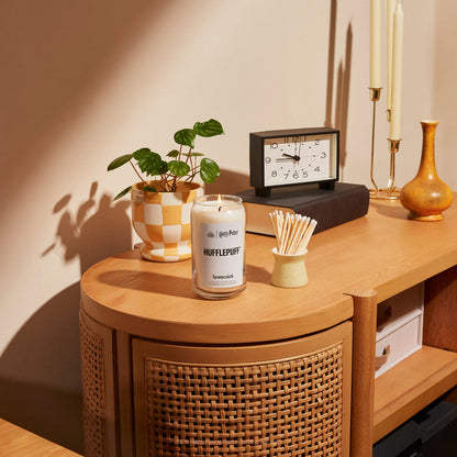 A lifestyle shot of a living room console table. On the table is the Hufflepuff candle next to a container of matches and other home decor items.
