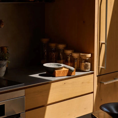 A warm, sunlit kitchen scene with light wood cabinetry, dark countertops, glass spice jars, and a small wooden mortar and pestle resting on the counter.
