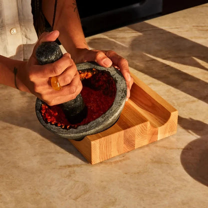 A person's hands using a dark gray stone mortar and pestle to grind red chili powder inside a light-colored wooden tray on a beige stone countertop.