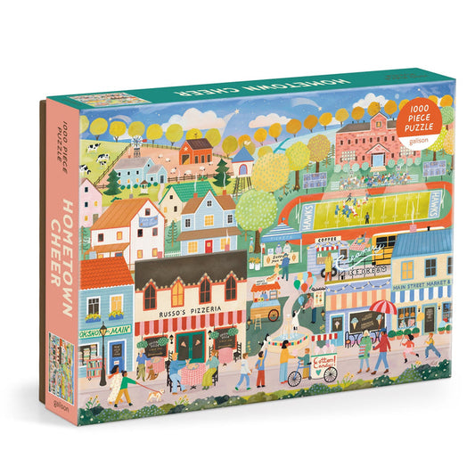 Hometown Cheer 1000 Piece Puzzle by Elisabeth Dorrian features a vibrant small town scene with shops, a pizzeria, market stalls, people strolling, trees, and houses.