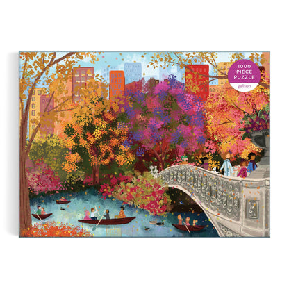 The Joy Laforme Autumn on Bow Bridge 1000 Piece Puzzle features a colorful illustration of people boating under Central Park’s Bow Bridge, framed by vibrant autumn trees and city buildings.