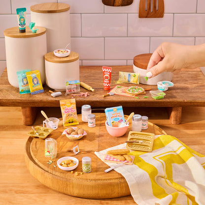 Miniature food items and kitchen accessories on a wooden surface with a hand interacting with them.