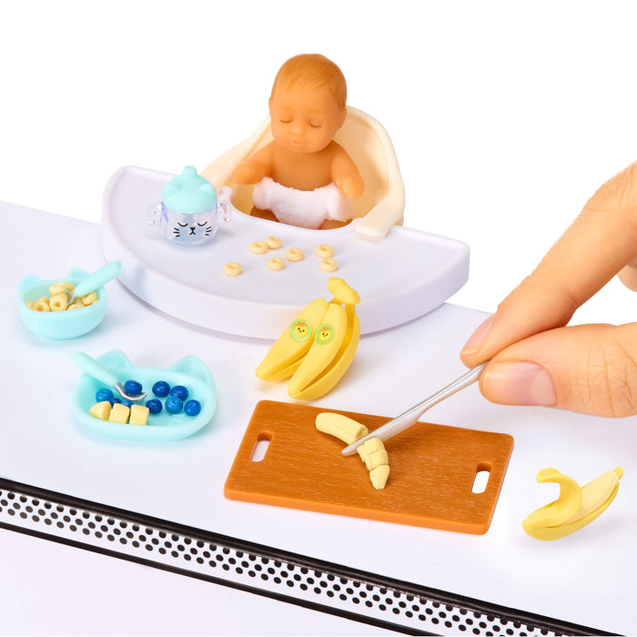 Miniature scene with a mini baby in a high chair, toys, and a hand holding a banana.