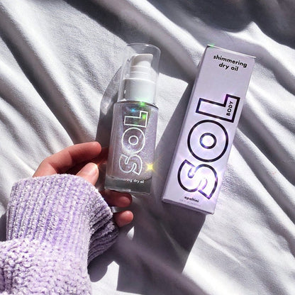 SOL Body Opaltini shimmering dry oil bottle with pump - sparkling with iridescent glitter - held in a hand beside its holographic SOL box on sunlit white bedding; ColourPop SOL Shimmering Dry Oil for a luminous body glow.